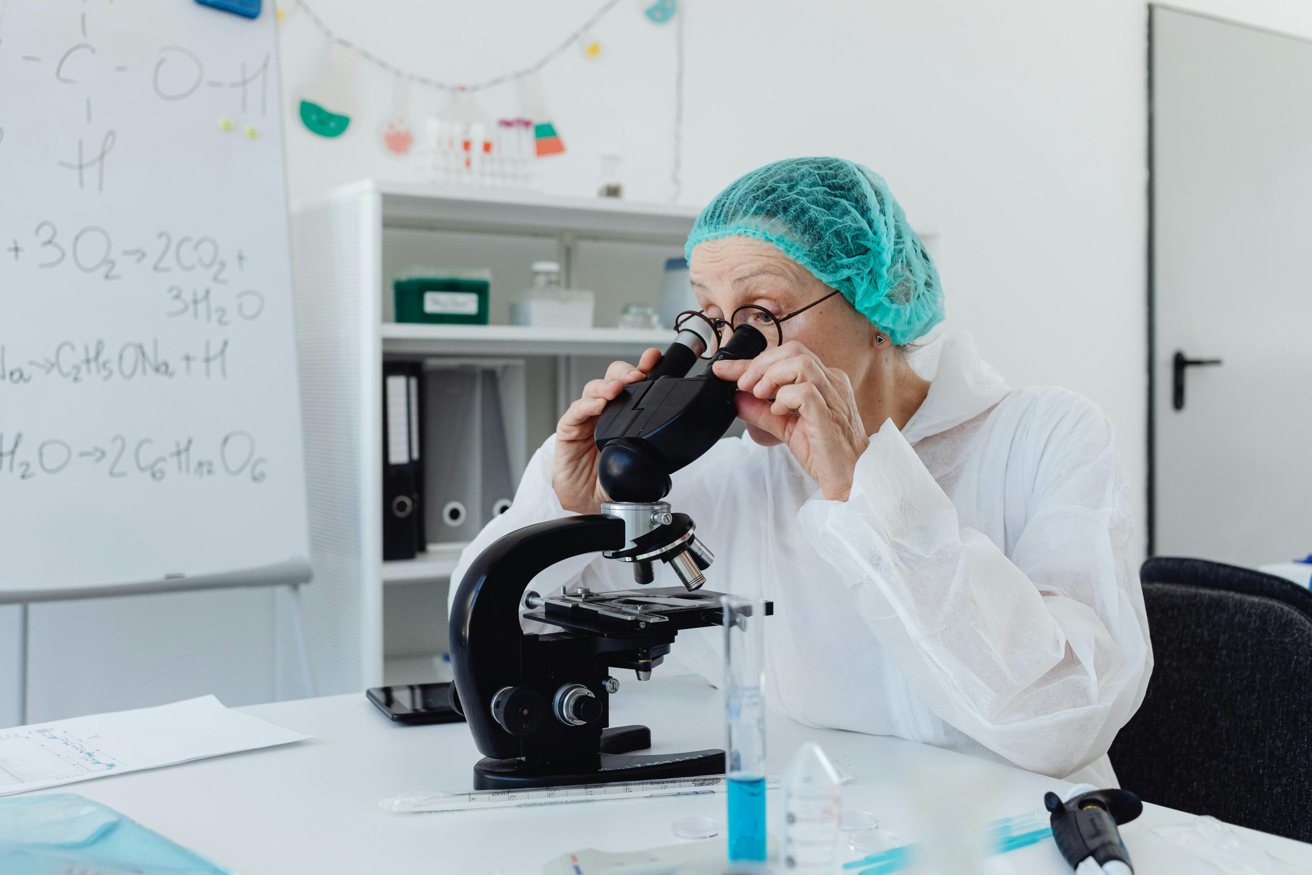 A senior female scientist using a microscope in a laboratory setting, wearing protective gear.
